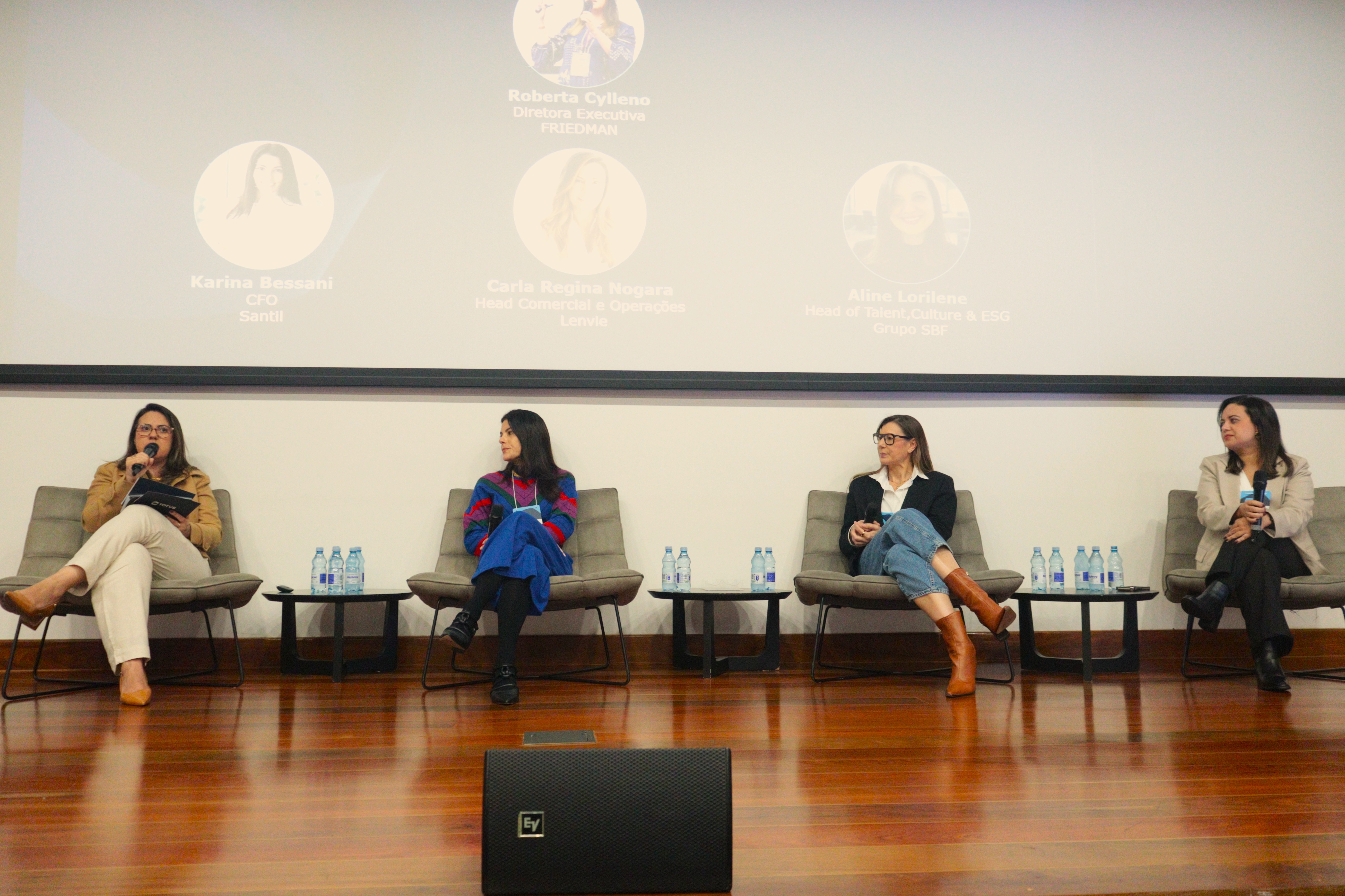 4 mulheres sentadas em poltronas, um painel com o nome e o cargo delas atrás.