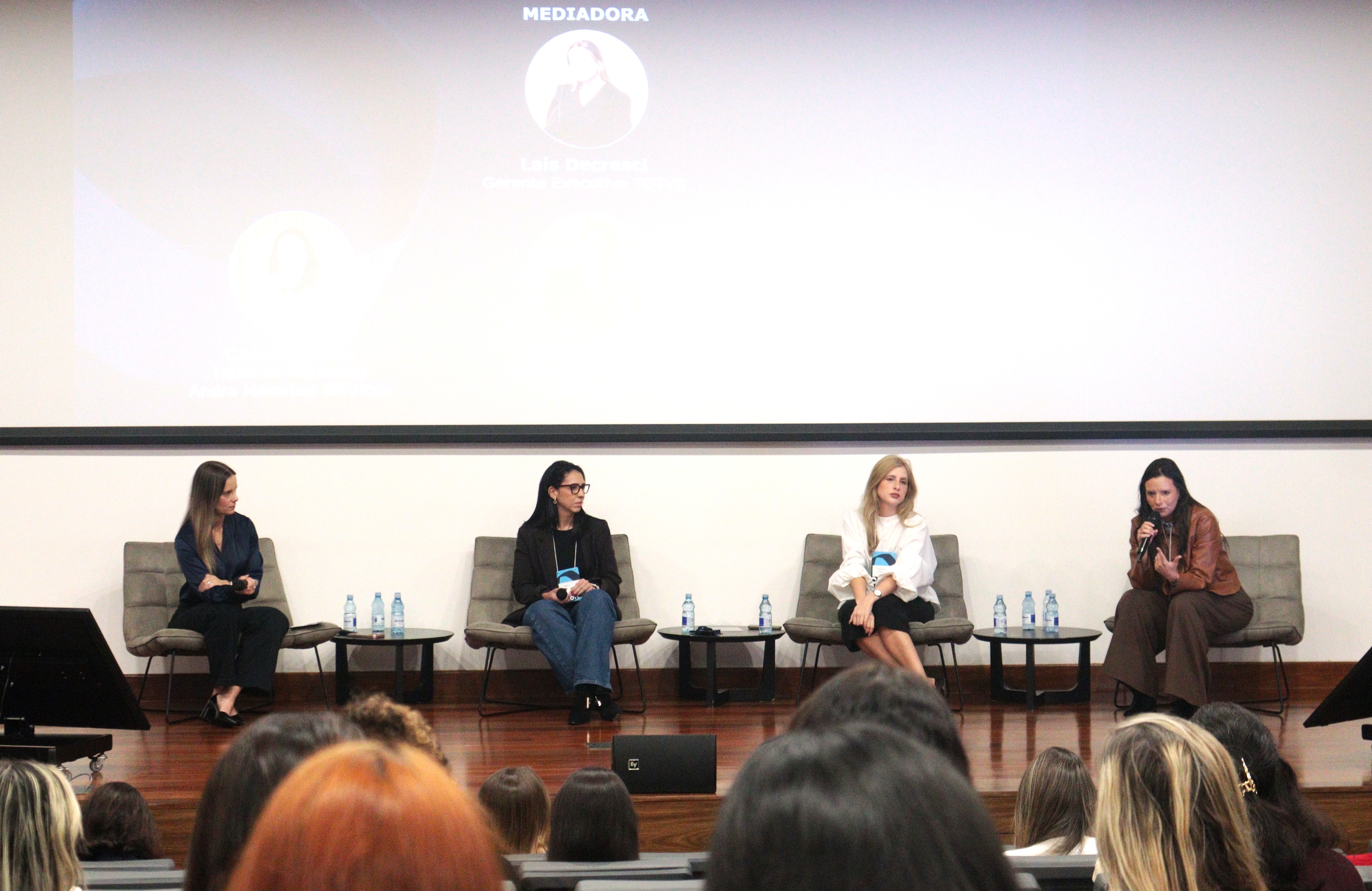 4 mulheres sentadas em poltronas, um painel com o nome e o cargo delas atrás.